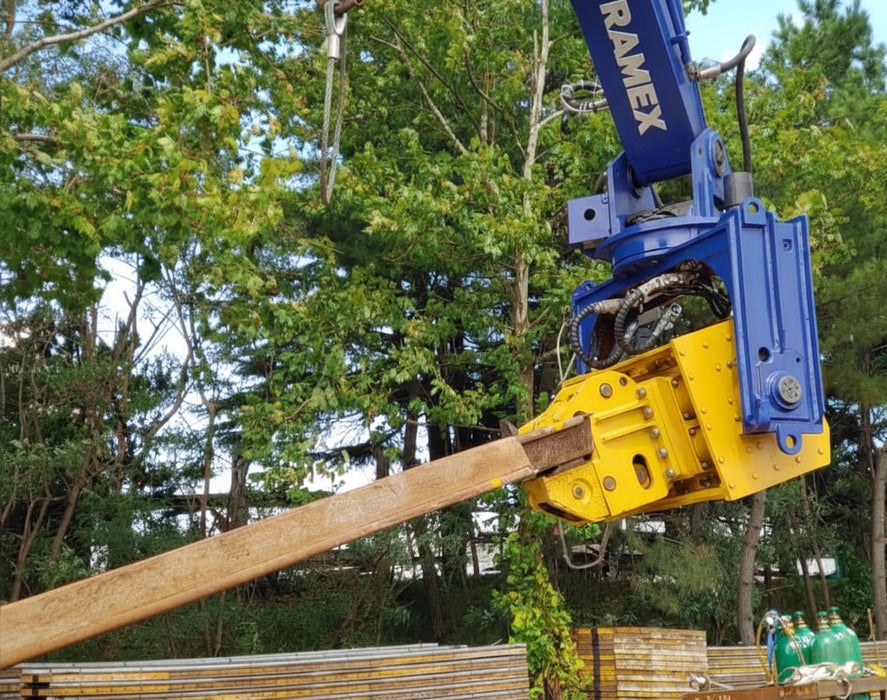 RAMEX auto-tilting vibro hammer gripping a steel pile at the edge of a wooded site